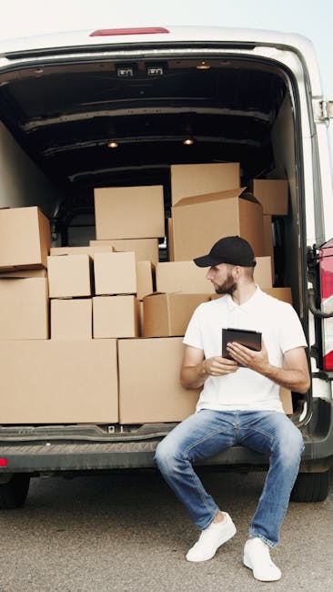 A man wearing a white t-shirt, blue jeans, and a black cap is sitting on the pavement next to an open van, which is filled with various packed cardboard boxes of different sizes, some sealed with brown packing tape. The boxes are arranged inside the vehicle, which appears to be used for home relocation purposes. The man is holding a tablet device, likely coordinating the packing or loading process. The background shows an outdoor setting, with the van positioned on a paved surface. This scene depicts a professional moving or removals service, such as those offered by Man with Van Merton, involved in furniture transport, packing, and loading during a house move, with equipment like packing materials and boxes visible to facilitate efficient relocation activities.