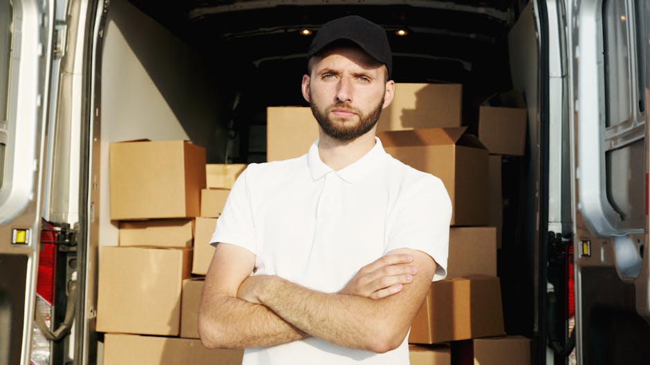A man wearing a white t-shirt, blue jeans, and a black cap is sitting on the pavement next to an open van, which is filled with various packed cardboard boxes of different sizes, some sealed with brown packing tape. The boxes are arranged inside the vehicle, which appears to be used for home relocation purposes. The man is holding a tablet device, likely coordinating the packing or loading process. The background shows an outdoor setting, with the van positioned on a paved surface. This scene depicts a professional moving or removals service, such as those offered by Man with Van Merton, involved in furniture transport, packing, and loading during a house move, with equipment like packing materials and boxes visible to facilitate efficient relocation activities.