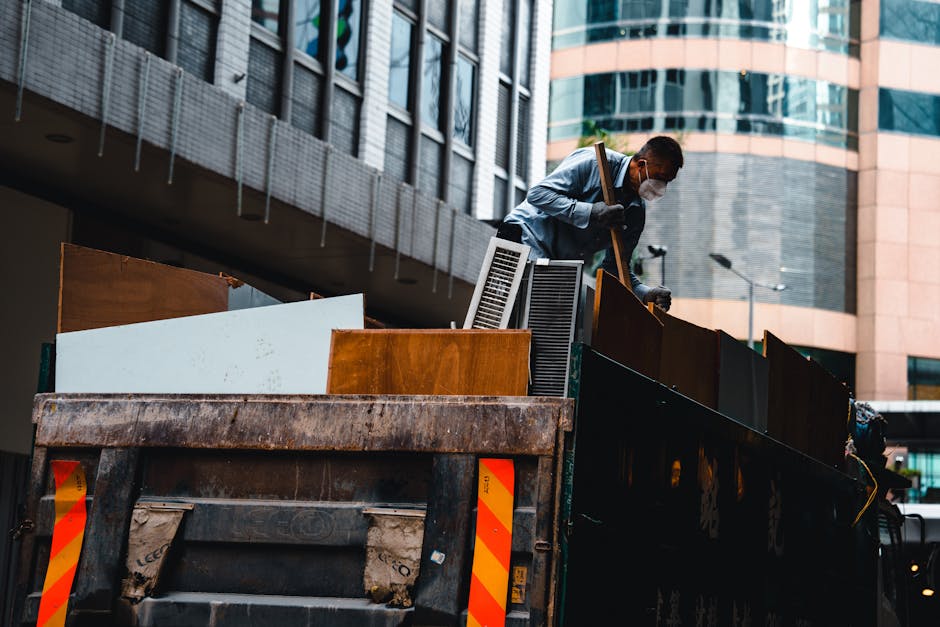 A man wearing a face mask and casual clothing is standing on the back of a black flatbed truck with orange and yellow reflective markings, loading household furniture and waste materials. The truck contains various furniture pieces, including wooden panels, a radiator, plastic containers, and cardboard boxes, all positioned towards the edge of the vehicle. The man is using a long-handled tool, possibly a broom or pry bar, to assist in the loading or organizing process of items intended for waste removal or house clearance. The scene takes place outdoors in an urban area with modern office buildings featuring glass and brick facades in the background. Soft natural lighting illuminates the scene, highlighting the textures of the furniture, materials, and the metallic surface of the truck. This image reflects the process of home relocation or furniture transport as part of a professional removals service, with a focus on proper disposal of bulky waste, aligned with the services offered by Man with Van Merton.