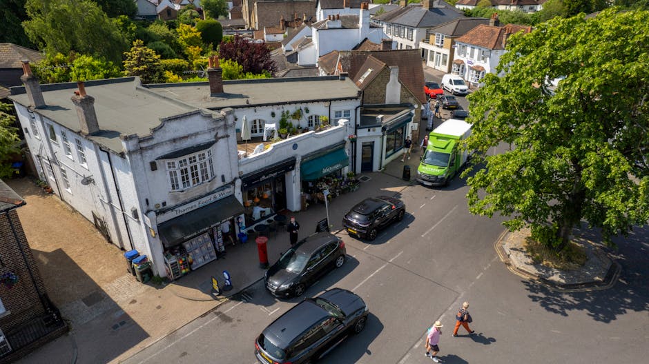 An aerial view of a street in Colliers Wood with a two-storey white building featuring large storefront windows and a dark awning, where Man with Van Merton is providing home relocation services. The building's entrance is open, and there are cardboard boxes and packing materials on the sidewalk in front. A black sedan and a grey estate car are parked directly outside, with a green van and a white delivery vehicle parked nearby. Two people are walking across the street while another individual is seen lifting or carrying boxes near the storefront, indicating an active loading process. A large, leafy green tree provides shade on one side of the street, and additional residential buildings and parked cars are visible further back, complementing the setting of a busy urban area engaged in furniture transport and packing activities during a moving operation.