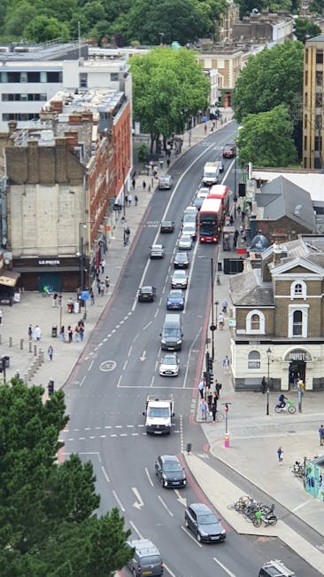 An aerial view of a busy street in Colliers Wood, featuring parked and moving vehicles including cars, vans, and a red double-decker bus along a curving road. On the left side, a narrow sidewalk is visible with pedestrians walking, some carrying bags or talking. The area is lined with a mix of older brick buildings and modern structures, with a row of large, leafy trees providing greenery along the road. On the right side, a small Victorian-style building with decorative trim and a windowed façade is situated next to bicycles parked at a rack. In the background, more trees and additional urban buildings are visible, emphasizing the lively, residential and commercial mix of the neighbourhood, relevant to home relocation and furniture transport services, such as those provided by Man with Van Merton.