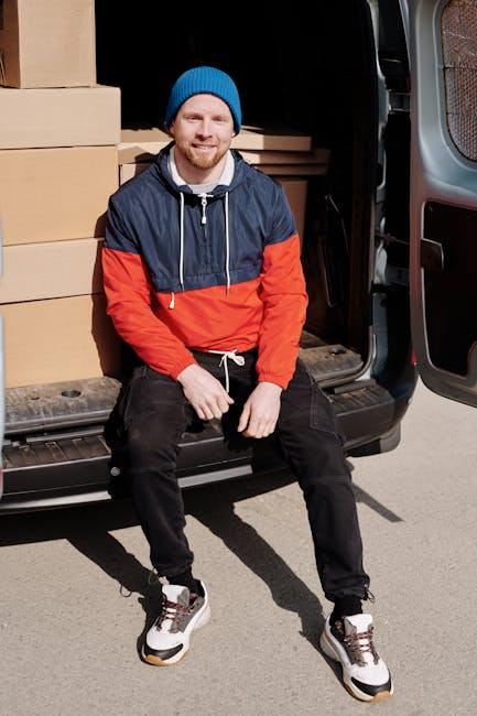 A man wearing a grey beanie, navy blue tracksuit, and a green padded vest is sitting on the edge of an open van during a home relocation process. Inside the vehicle, several cardboard boxes of various