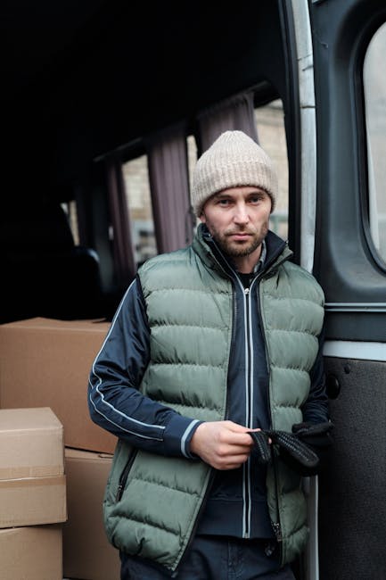 A man with a serious expression, wearing a beige knitted beanie and a padded green and navy jacket, stands inside a vehicle with a black interior. Behind him, there are cardboard boxes stacked neatly, suggesting a home relocation or moving process. The vehicle appears to be a van used for furniture transport, with part of the door open revealing the loading area. The image is well-lit, highlighting the details of the man's clothing, the cardboard packaging materials, and the interior environment, indicating a professional move or packing and moving services by Man with Van Merton involved in emergency same-day removals in Merton.