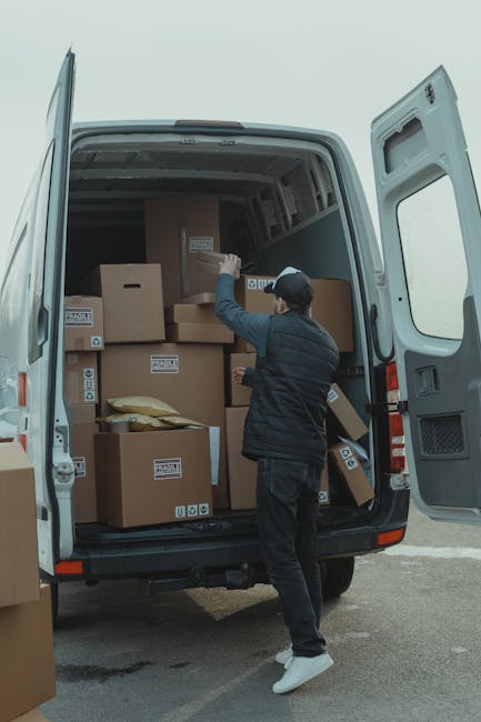A person wearing a dark jacket, grey trousers, and a cap is standing next to the open rear doors of a large white van, which is parked outdoors on a paved area. Inside the van, numerous cardboard boxes of various sizes are stacked, some sealed with packing tape and labelled with barcodes or handling instructions. The boxes are arranged to maximize space and facilitate efficient loading or unloading during a home relocation, with some cushions or padded materials visible among the boxes. The individual appears to be in the process of placing or retrieving a box, using their right hand to gesture towards the interior. Equipment such as a small trolley or straps is not visible, but the scene clearly depicts the loading process common in furniture transport and packing operations by [COMPANY_NAME], which offers removals services to assist with house moves, ensuring compliance with local regulations and minimizing legal risks related to loading bays and parking restrictions in Merton. The background shows an overcast sky, indicating typical outdoor conditions during a home move or furniture transport task.