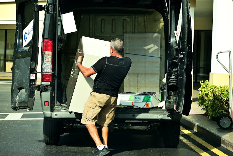 A man with short gray hair, dressed in a black t-shirt and beige shorts, is loading a large white cabinet into the back of a black van parked on a paved street outside a modern building. The van's rear doors are open, revealing a spacious interior with additional furniture and packing materials, including cardboard boxes and plastic-wrapped items. The loading area is adjacent to a sidewalk with a small green bush and a wheeled luggage cart nearby. The scene is illuminated by daylight, emphasizing the process of home relocation and furniture transport, as Man with Van Merton facilitates packing and loading activities for house removals, with the van positioned close to the building's entrance.