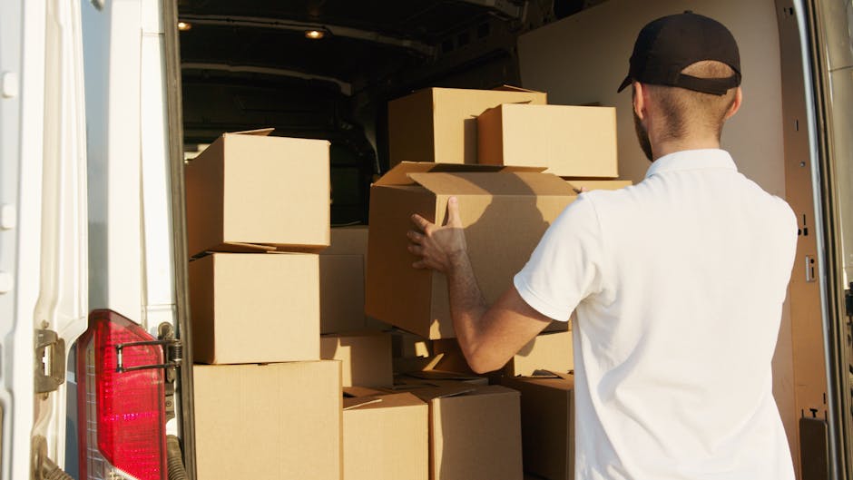 A person wearing a dark jacket, grey trousers, and a cap is standing next to the open rear doors of a large white van, which is parked outdoors on a paved area. Inside the van, numerous cardboard boxes of various sizes are stacked, some sealed with packing tape and labelled with barcodes or handling instructions. The boxes are arranged to maximize space and facilitate efficient loading or unloading during a home relocation, with some cushions or padded materials visible among the boxes. The individual appears to be in the process of placing or retrieving a box, using their right hand to gesture towards the interior. Equipment such as a small trolley or straps is not visible, but the scene clearly depicts the loading process common in furniture transport and packing operations by [COMPANY_NAME], which offers removals services to assist with house moves, ensuring compliance with local regulations and minimizing legal risks related to loading bays and parking restrictions in Merton. The background shows an overcast sky, indicating typical outdoor conditions during a home move or furniture transport task.