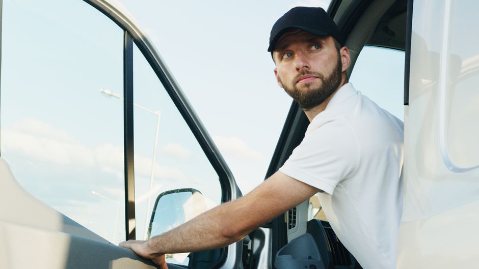 A male mover with a beard, wearing a black cap and white t-shirt, is inside the cab of a white moving van, looking outward with a serious expression. His right arm is extended forward, resting on the dashboard, as he assists with the home relocation process. The interior of the van is visible behind him, with the windshield and side window providing a view of a cloudy sky. Outside the van, part of the pavement and a parking area can be seen, suggesting he is preparing for furniture transport or loading inside a property. The scene, captured during daytime, reflects the logistics involved in packing and moving, with a focus on vehicle loading preparations essential for efficient removals. Man with Van Merton often handles such loading processes, ensuring careful handling of boxes, furniture, and appliances during home removals and furniture transport services.