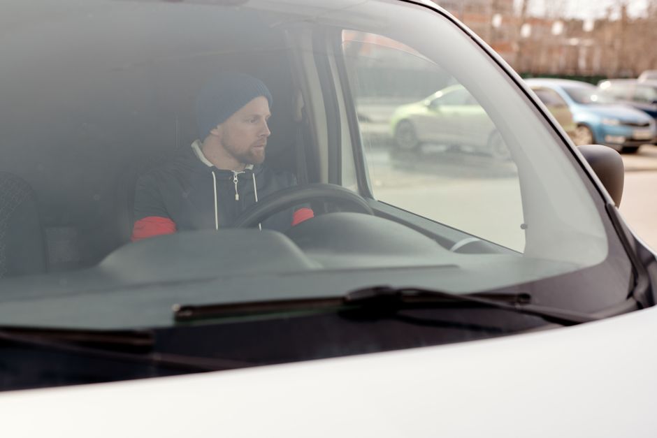 Close-up view through the front windshield of a vehicle showing a man with a beard and wearing a dark beanie, black jacket with red accents, sitting in the driver's seat. Inside the vehicle, the dashboard and steering wheel are visible, and in the background, parked cars and a residential or commercial area outdoors can be seen. This scene captures the process of a home relocation or furniture transport, possibly part of a loading or move planning stage, with the vehicle used by Man with Van Merton during a removals service, indicated by the context and website details.
