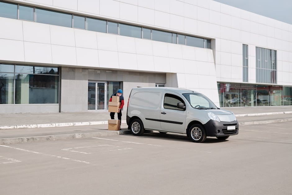 A person dressed in a blue hat and red jacket is standing beside a small, light grey commercial van parked in an empty outdoor parking area with marked spaces. The individual is holding and stacking cardboard boxes, preparing for a home relocation or furniture transport process. The van is positioned close to the curb in front of a modern building with large glass windows and white exterior panels. The scene appears to be during daylight hours with clear or overcast weather, facilitating a loading or unloading activity related to house removals. Man with Van Merton, a specialist in removals services, may be involved in this logistics operation, using the van and cardboard boxes for packing and moving furniture or household items.
