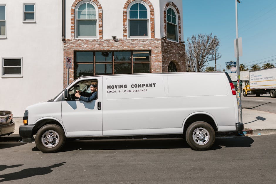 A white moving van with the company name 'MOVING COMPANY' and the tagline 'LOCAL & LONG DISTANCE' printed on the side is parked beside a brick building with large, arched windows. The vehicle is on an urban street with other parked cars and trucks visible in the background. A man wearing sunglasses and a jacket is sitting in the driver's seat, leaning out of the open window, possibly preparing for a home relocation or furniture transport. The scene is set during daylight hours with clear weather, and the pavement beneath the van displays a no-parking sign attached to the building. The surroundings include a sidewalk, street signs, and a few trees, indicating the vehicle is positioned in a city environment suitable for removals services, as provided by Man with Van Merton, specializing in house removals, packing, and moving logistics.
