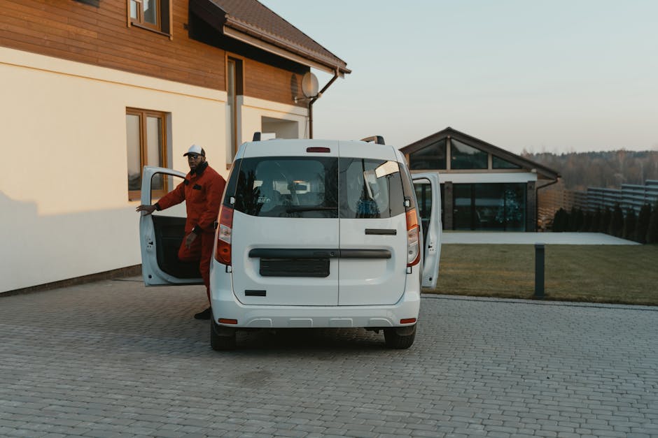 A person dressed in a blue hat and red jacket is standing beside a small, light grey commercial van parked in an empty outdoor parking area with marked spaces. The individual is holding and stacking cardboard boxes, preparing for a home relocation or furniture transport process. The van is positioned close to the curb in front of a modern building with large glass windows and white exterior panels. The scene appears to be during daylight hours with clear or overcast weather, facilitating a loading or unloading activity related to house removals. Man with Van Merton, a specialist in removals services, may be involved in this logistics operation, using the van and cardboard boxes for packing and moving furniture or household items.