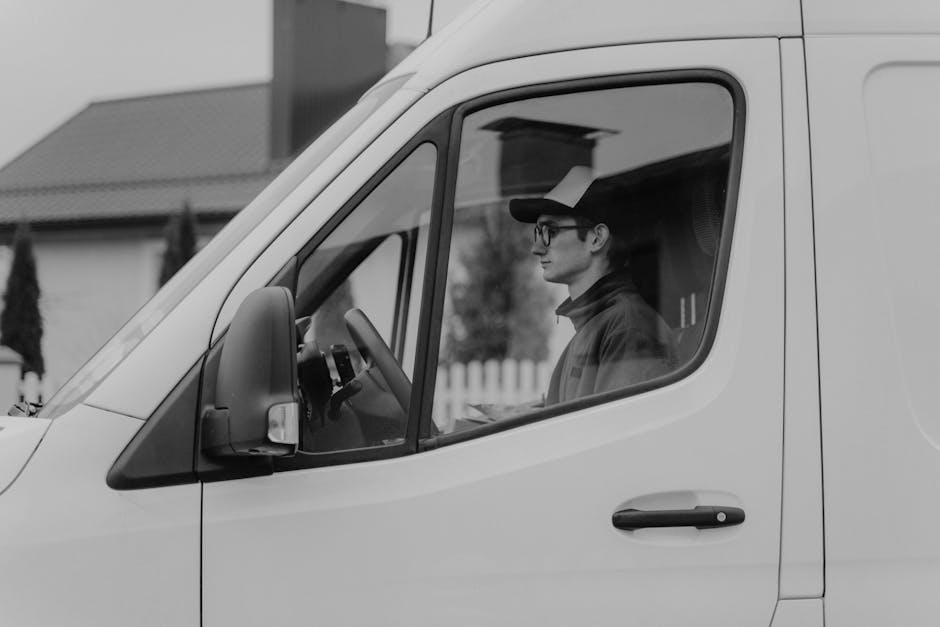 A man with short hair, wearing glasses, a dark cap, and a dark jacket, is seated inside the cab of a white delivery van, with his side visible through the window. The van is parked on a residential street, evident from the background which features houses with tiled roofs, a white picket fence, and some trees. The interior of the van's cab shows the steering wheel, dashboard, and part of the side mirror. The man appears focused, possibly preparing for a home relocation or furniture transport, aligning with the services offered by Man with Van Merton for removals. The scene captures the loading process or vehicle preparation stage, with the vehicle close to the property for easy access to load or unload items like cardboard boxes, wrapped furniture, and packing materials, which are typically used during packing and moving operations in house removals.