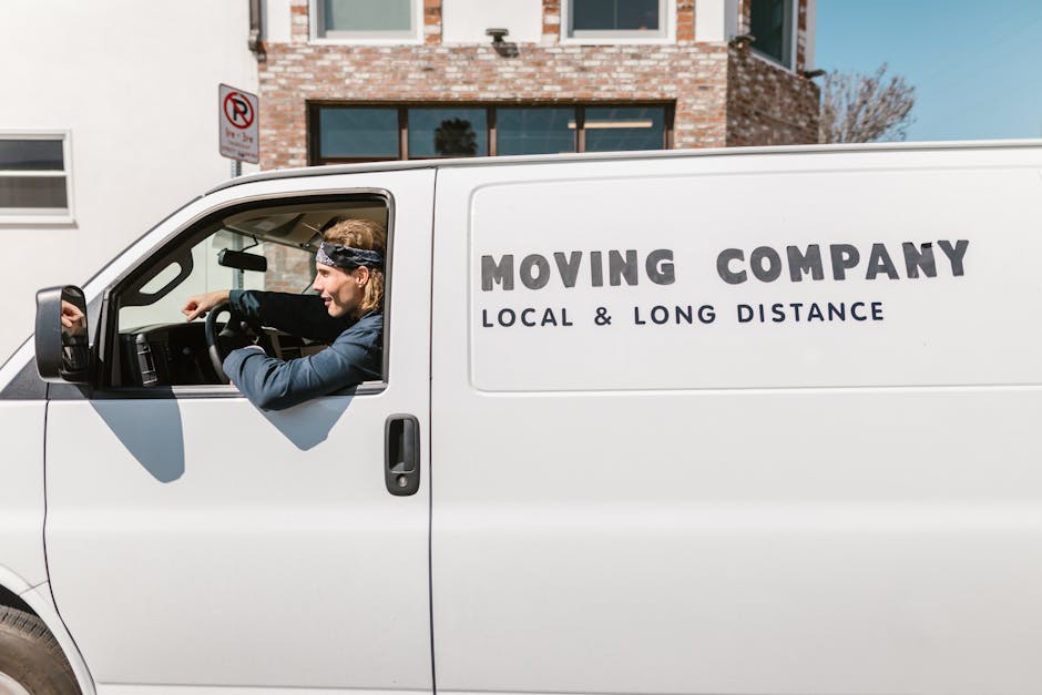 A white moving van with the text 'Moving Company Local & Long Distance' printed on its side is parked outside a residential building with brick and white exterior walls and large windows. Inside the driver’s cabin, a person with blonde hair, wearing a dark jacket and a patterned headband, is seen reaching out and preparing to load or unload furniture or boxes. The scene suggests a home relocation process, with the individual involved in the logistics of packing and furniture transport. The van is positioned on a paved driveway or street, close to the entrance of the property, which has an open doorway possibly indicating active loading. The environment is well-lit, with clear weather, and the scene exemplifies professional moving services connected to house removals near Wimbledon Common, supported by Man with Van Merton.