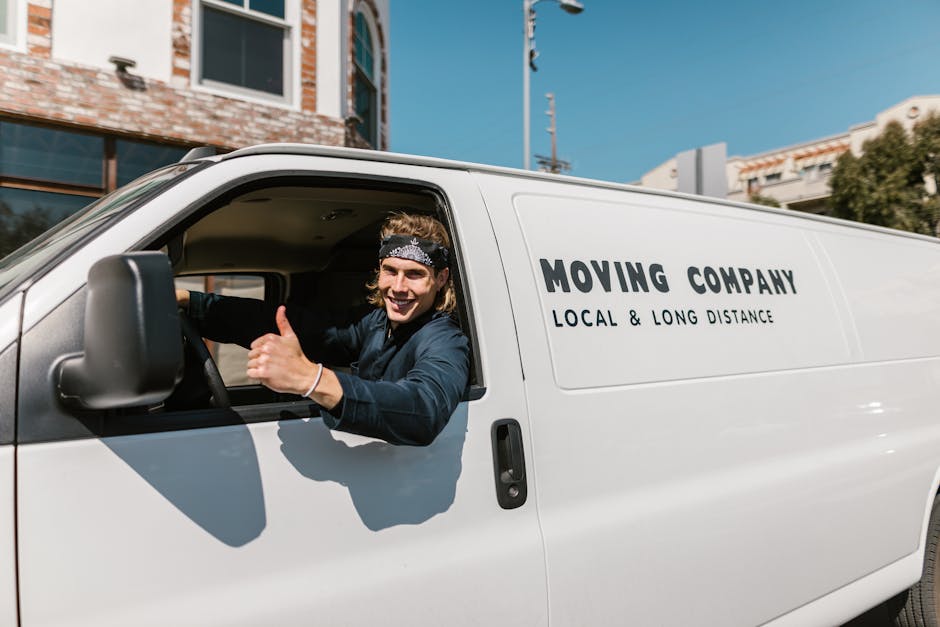 A smiling male mover with long hair, wearing a black bandana and dark clothing, is sitting in the driver's seat of a white van with 'MOVING COMPANY, LOCAL & LONG DISTANCE' printed on the side, giving a thumbs-up gesture. The van is parked outside a residential building with a brick and glass facade, and nearby is a clear blue sky. The vehicle appears ready for a home relocation or furniture transport, with the driver preparing for a packing and moving service. Visible in the background are street signs, electrical poles, and some greenery, indicating an urban neighbourhood environment typical for house removals near Wimbledon Common. Man with Van Merton’s services involve loading and unloading furniture and boxes, and the scene captures a typical moment during the loading process or before commencing a house move.