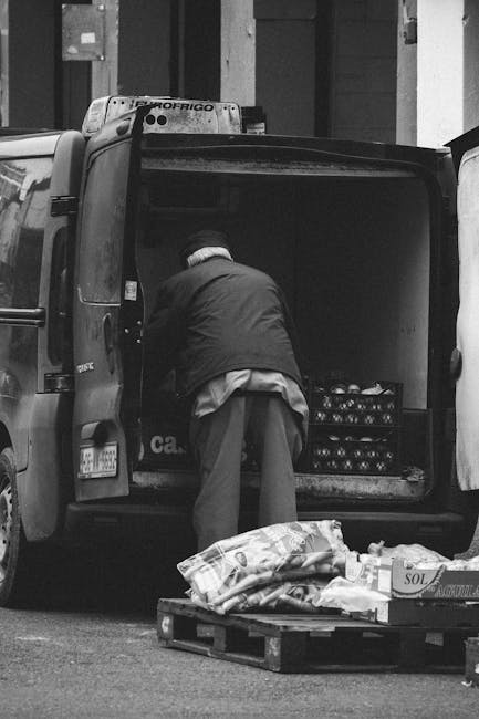A man with a hat and dark jacket is loading the rear of a black van, parked on a street in front of a building, with a crate of bottles visible inside. The van is part of a home relocation or furniture transport process carried out by Man with Van Merton, a professional removals service. In front of the van, there is a wooden pallet with wrapped and covered items, including fabric and cardboard packaging materials, which are likely household goods being packed for moving. The scene appears during daylight, revealing urban surroundings with other parked vehicles nearby. The van's open tailgate shows the interior being prepared for loading additional items as part of a packing and moving operation. The environment emphasizes the logistics involved in residential removals, including careful handling of boxes and furniture, supported by professional moving equipment like pallets and straps.