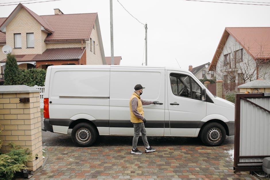 A man wearing a yellow and brown jacket, black cap, and face mask is standing outside a residential property, opening the side sliding door of a white panel van parked on a paved driveway. The van, used for home relocation, has its rear and side doors closed. Nearby, a brick wall and garden fencing frame the scene, with a row of houses featuring pitched roofs and brick exteriors visible in the background. The scene appears to be during daytime with overcast weather, and the environment suggests the initial stages of packing and furniture transport, a typical part of the loading process handled by a professional removals service such as Man with Van Merton.