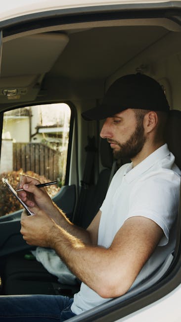 A man with a hat and dark jacket is loading the rear of a black van, parked on a street in front of a building, with a crate of bottles visible inside. The van is part of a home relocation or furniture transport process carried out by Man with Van Merton, a professional removals service. In front of the van, there is a wooden pallet with wrapped and covered items, including fabric and cardboard packaging materials, which are likely household goods being packed for moving. The scene appears during daylight, revealing urban surroundings with other parked vehicles nearby. The van's open tailgate shows the interior being prepared for loading additional items as part of a packing and moving operation. The environment emphasizes the logistics involved in residential removals, including careful handling of boxes and furniture, supported by professional moving equipment like pallets and straps.