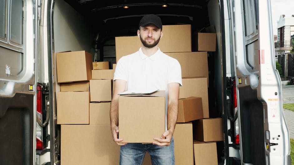 A man wearing a white polo shirt and cap is standing outside the back of a van during a home relocation, holding a cardboard box as part of a furniture transport process. The van's interior is visible